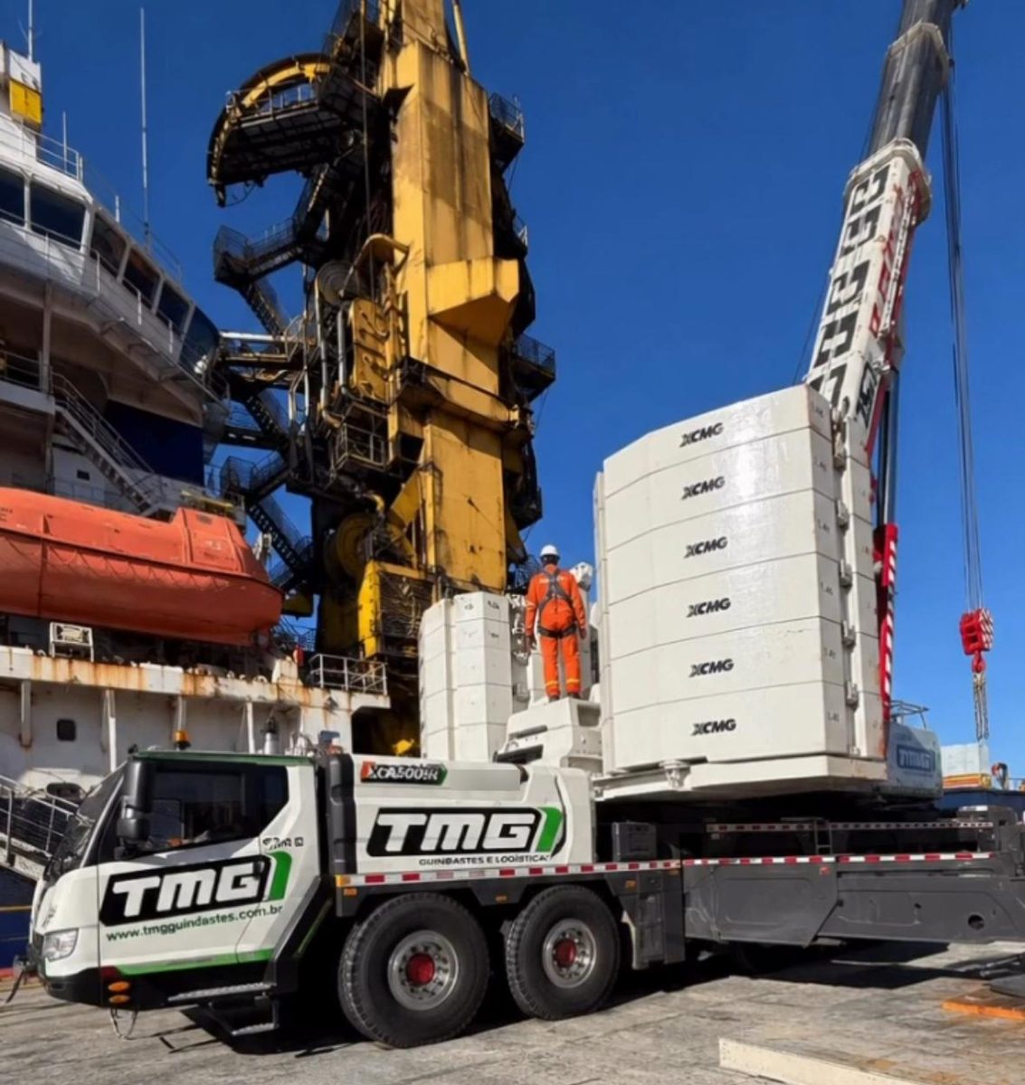 Guindaste rodoviário com contrapesos montados ao lado de um navio; trabalhador de capacete, macacão e cinto sobre a máquina